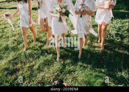 Les demoiselles et les épouses sans visage vêtues de robes de satin avec de beaux bouquets dans leurs mains marchent pieds nus sur l'herbe verte dans le jardin. Matin Banque D'Images