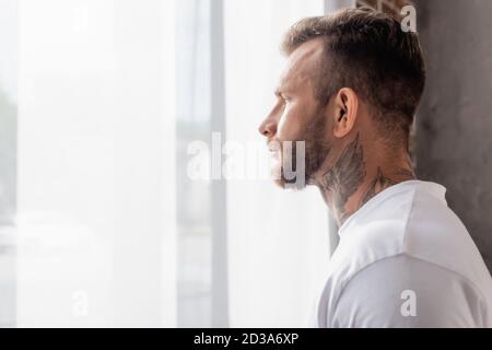 vue latérale de l'homme barbu, tatoué, regardant loin en se tenant près de la fenêtre à la maison Banque D'Images