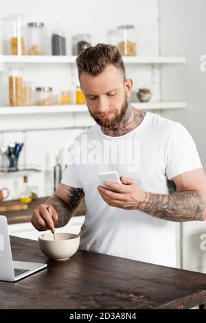 un indépendant à barbe et tatoué mélangeant le petit-déjeuner tout en discutant sur un téléphone portable dans la cuisine Banque D'Images