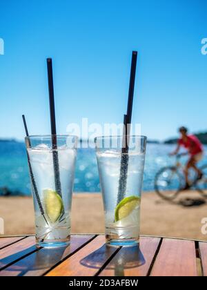 Deux verres de gin tonique avec pailles noires sur un bureau en bois. Mer et motard sur fond flou. Banque D'Images