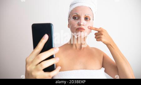 Une femme caucasienne enveloppée dans des serviettes blanches avec un masque cosmétique prend selfie avec un téléphone portable. Banque D'Images