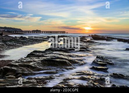 Le lever du soleil d'octobre derrière la jetée Hastings dans l'est de Sussex et La marée descendante révélant les rochers sur la plage de St Leonards Banque D'Images