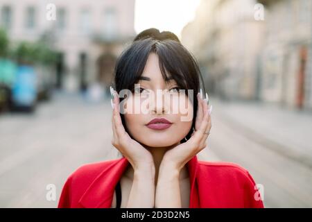 Gros plan visage portrait extérieur de la jolie brunette asiatique modèle féminin avec des cheveux noirs et maquillage de mode, penchée visage sur ses paumes, posant à l'appareil photo Banque D'Images