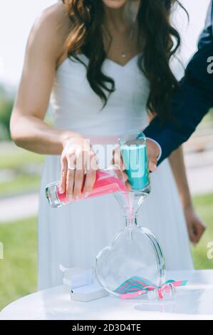 Gros plan de la mariée et du marié versant du sable rose et bleu dans un vase en verre en forme de coeur pendant la célébration de son mariage. Salon de mariage Banque D'Images