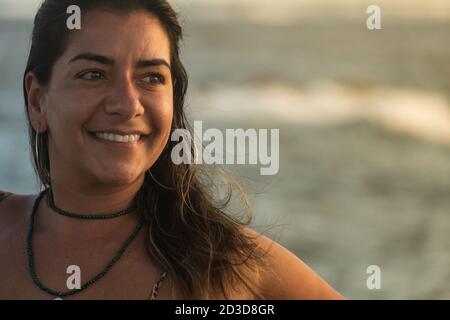 Belle femme brune sourit illuminée par la lumière du coucher de soleil avec la mer et les vagues en arrière-plan. Banque D'Images
