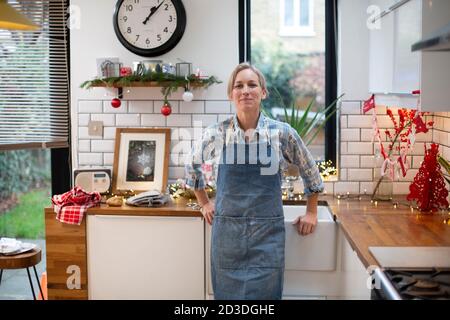 Femme blonde portant un tablier bleu debout dans la cuisine, souriant à la caméra. Banque D'Images