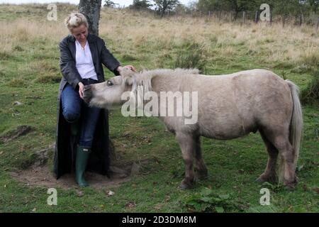 Blanche blanche blanche blanche teint blonde moyenne d'âge femme dans ses 40 ans portant un long manteau de cuir acheté d'une deuxième main boutique dans le champ avec des chevaux et des poneys. Banque D'Images