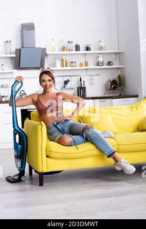 Femme au foyer avec la main sur la hanche assis sur le canapé et tenant aspirateur à la maison Banque D'Images
