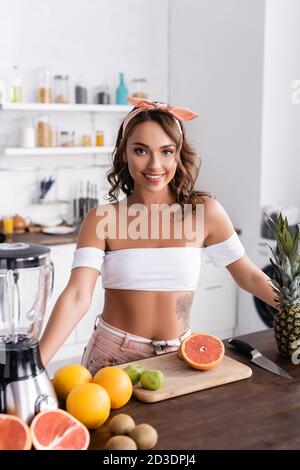 Mise au point sélective de la femme brunette regardant la caméra près de mûr fruits et mixeur Banque D'Images