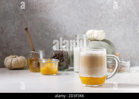 Verre de latte aux épices en couches de citrouille avec purée de citrouille, mousse de lait et cannelle debout avec ingrédients en pots et citrouilles blanches décoratives sur whi Banque D'Images