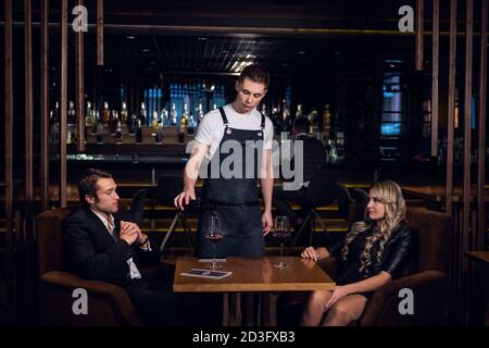 un serveur expérimenté dans un tablier verse du vin rouge dans des verres pour un jeune blond et un beau homme. Banque D'Images