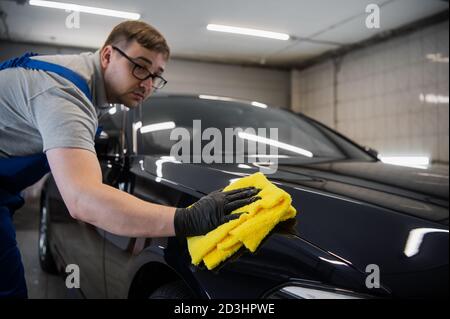 L'homme après le lavage essuie la voiture noire avec un chiffon au lavage de voiture. Main mâle et carrosserie de voiture de près. Banque D'Images