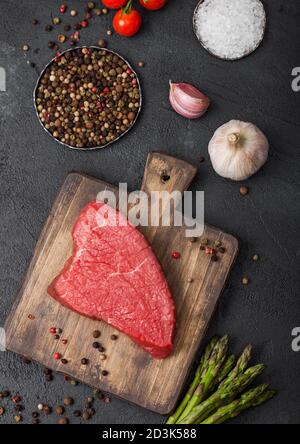 Tranche de steak de bœuf cru sur planche à découper en bois avec tomates, ail et pointes d'asperges sur fond sombre de table de cuisine. Banque D'Images