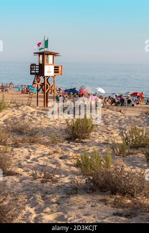Côte de plage avec beaucoup de sable et quelques plantes. Il y a une tour de maître-nageur avec croix rouge et drapeaux verts. Sur la plage il y a des gens en Banque D'Images