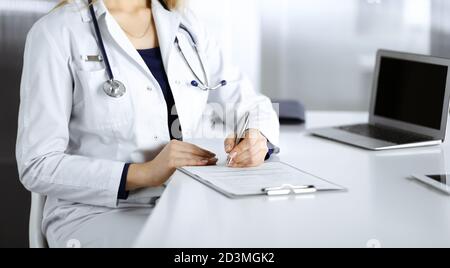 Une femme-médecin inconnue prescrit des médicaments à son patient, à l'aide d'un presse-papiers, tout en étant assise sur le bureau de son cabinet. Femme médecin Banque D'Images