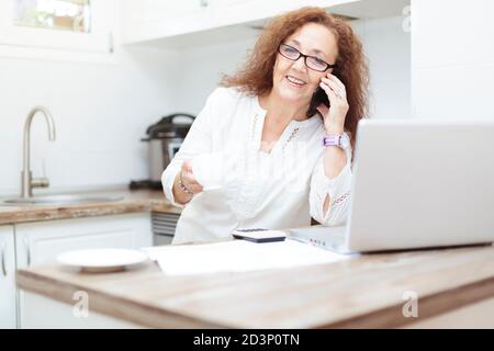Une femme de 60 ans utilisant son smartphone dans la cuisine. Elle est debout à côté d'un ordinateur portable et certains documents sont répartis sur la table. Banque D'Images
