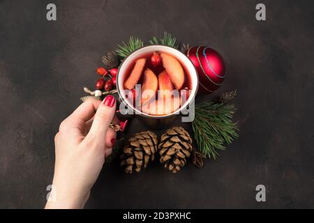 Composition de Noël, une main de femme met une tasse au centre des jouets de Noël et des branches de pin sur un fond de béton brun. Banque D'Images