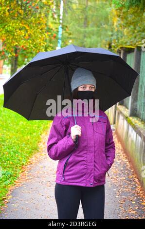 Jeune femme en vêtements chauds, avec masque noir réutilisable et chapeau en tricot. Tenir un parapluie ouvert debout sur le trottoir recouvert de feuilles. Automne. Banque D'Images