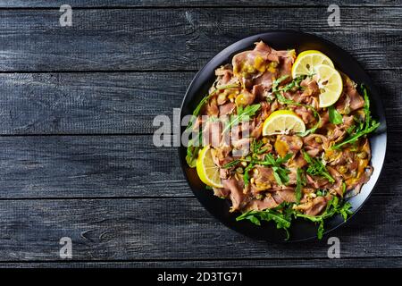 salade de thon finement tranché avec câpres, roquette, pignons et vinaigrette à la moutarde sur une assiette noire sur une table en bois, vue horizontale depuis le dessus, plat, Banque D'Images