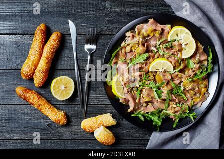 salade de thon finement tranché avec câpres, roquette, pignons et vinaigrette à la moutarde sur une assiette noire sur une table en bois avec des petits pains au sésame, vue horizontale depuis Banque D'Images