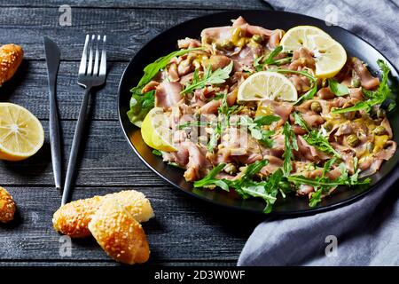 salade de thon finement tranché avec câpres, roquette, pignons et vinaigrette à la moutarde sur une assiette noire sur une table en bois avec des petits pains au sésame, vue horizontale depuis Banque D'Images