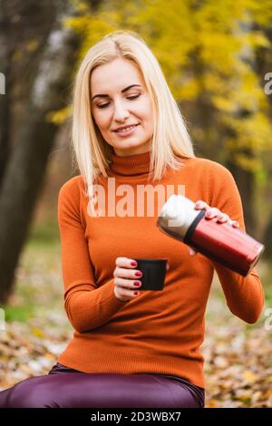 Jolie femme blonde dans des vêtements lumineux ouvrir métal thermos et verser le thé, le café dans la tasse à l'arrière-plan de l'automne Banque D'Images