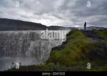 Un homme portant un manteau à capuchon debout sur des marches de pierre près du bord de la chute d'eau de Dettifoss dans le nord-est de l'Islande comme il plonge dans le canyon ci-dessous. Banque D'Images