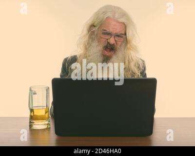 Studio shot of senior homme barbu en colère crier tout en utilisant un ordinateur portable avec verre de bière sur la table en bois Banque D'Images