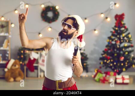 Homme en débardeur, chapeau de père Noël et lunettes de soleil drôles chantant et se moquant à la maison avec le micro à la main Banque D'Images