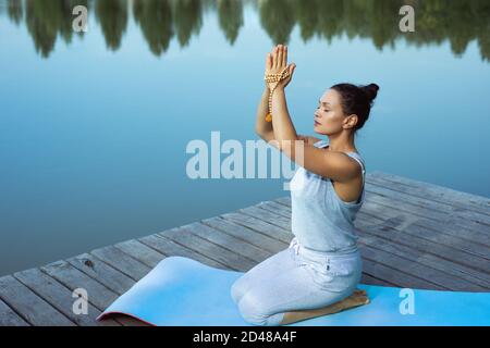 La jeune femme s'assied sur un tapis près du lac avec ses mains pliées soulevées au-dessus de sa tête. Méditation, yoga dans la nature. Banque D'Images