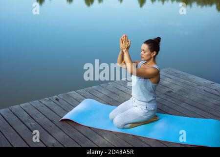 La jeune femme s'assied sur un tapis près du lac avec ses mains pliées soulevées au-dessus de sa tête. Méditation, yoga dans la nature. Banque D'Images