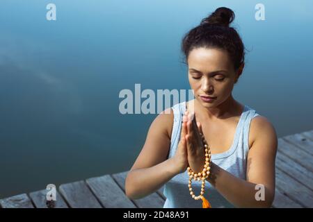 La jeune femme s'assied sur un tapis près du lac en tenant ses mains pliées devant elle. Méditation, yoga dans la nature. Banque D'Images