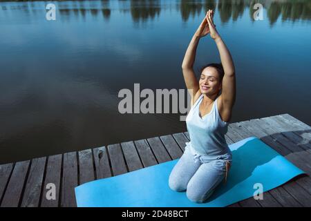 La jeune femme s'assoit sur un tapis bleu sur un quai en bois et étire ses bras vers le haut. Méditation, yoga dans la nature. Banque D'Images