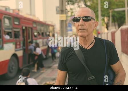 Senior homme chauve touristique pensée tout en portant des lunettes de soleil à l'arrêt de bus à Bangkok, Thaïlande Banque D'Images