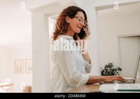 Femme heureuse travaillant sur ordinateur portable et parlant sur téléphone portable à la maison. femme d'affaires assise à table et travaillant. Banque D'Images