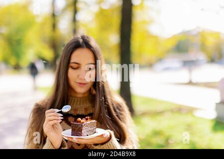 Souriante mannequin caucasienne mangeant un gâteau de tranche à l'extérieur sur fond de parc vert, appréciant le dessert au chocolat doux et le café, fille manger Banque D'Images