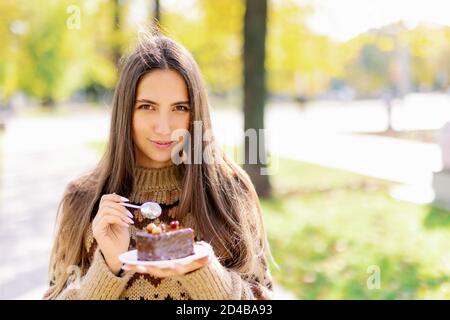 Souriante mannequin caucasienne mangeant un gâteau de tranche à l'extérieur sur fond de parc vert, appréciant le dessert au chocolat doux et le café, fille manger Banque D'Images