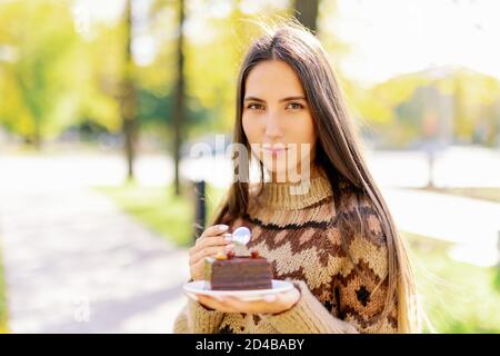 Souriante mannequin caucasienne mangeant un gâteau de tranche à l'extérieur sur fond de parc vert, appréciant le dessert au chocolat doux et le café, fille manger Banque D'Images