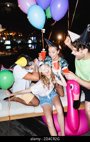 Objectif sélectif de la femme tenant une tasse jetable près des amis dans casquettes et ballons de fête la nuit Banque D'Images