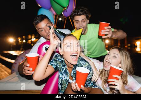 Objectif sélectif d'une femme excitée portant des casquettes de fête près d'amis avec des tasses jetables et des ballons à l'extérieur la nuit Banque D'Images