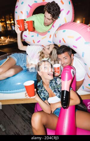 Attention sélective de la femme excitée assise sur l'anneau de natation près petit ami et amis avec des tasses jetables la nuit Banque D'Images