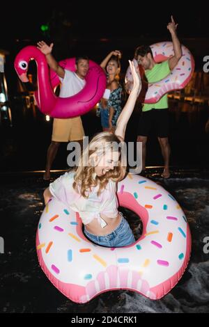 Objectif sélectif de la femme excitée avec anneau de natation debout piscine près des amis la nuit Banque D'Images