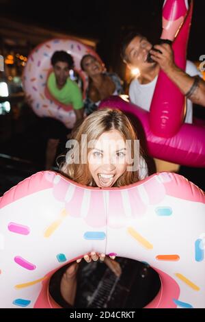 Objectif sélectif de la femme excitée avec anneau de natation regardant caméra près de vos amis la nuit Banque D'Images