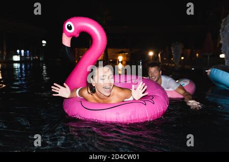 Objectif sélectif de la femme excitée nageant en anneau dans la piscine proche petit ami la nuit Banque D'Images