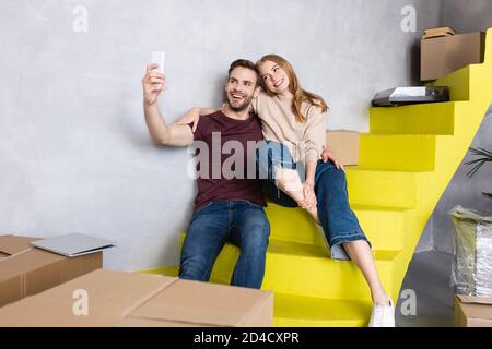 un couple excité assis sur des escaliers jaunes et prenant le selfie à proximité boîtes en carton Banque D'Images