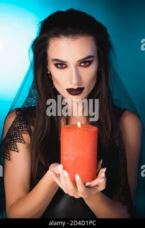 femme pâle avec maquillage noir et voile tenant une bougie brûlante sur bleu Banque D'Images