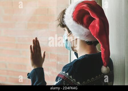 Enfant avec masque médical portant chapeau de Père Noël attendez le père noël à côté d'une fenêtre à Noël par le Pandémie de coronavirus Banque D'Images