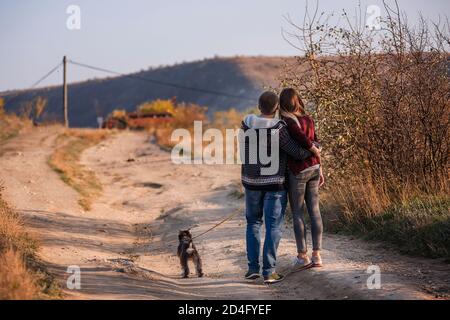 Une jeune famille heureuse marche avec un chien Schnauzer le long de la route, se tenant les mains, regardant l'une l'autre sur un fond de ciel bleu et de forêt. Banque D'Images