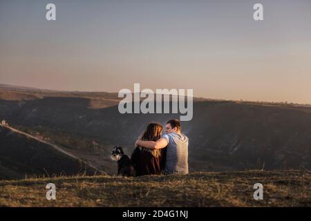 Un couple heureux amoureux se repose sur le flanc de la montagne avec un Schnauzer noir, assis sur le sol, embrassant un chien, regardant les parapentes lumineuses voler Banque D'Images