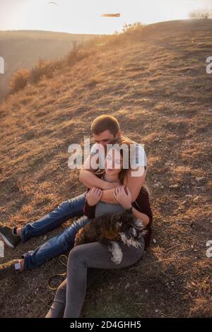 Un jeune homme embrasse une belle fille de derrière, le couple admirer le coucher du soleil depuis le sommet de la colline. Une famille heureuse voyage avec un chien schnauzer. Banque D'Images